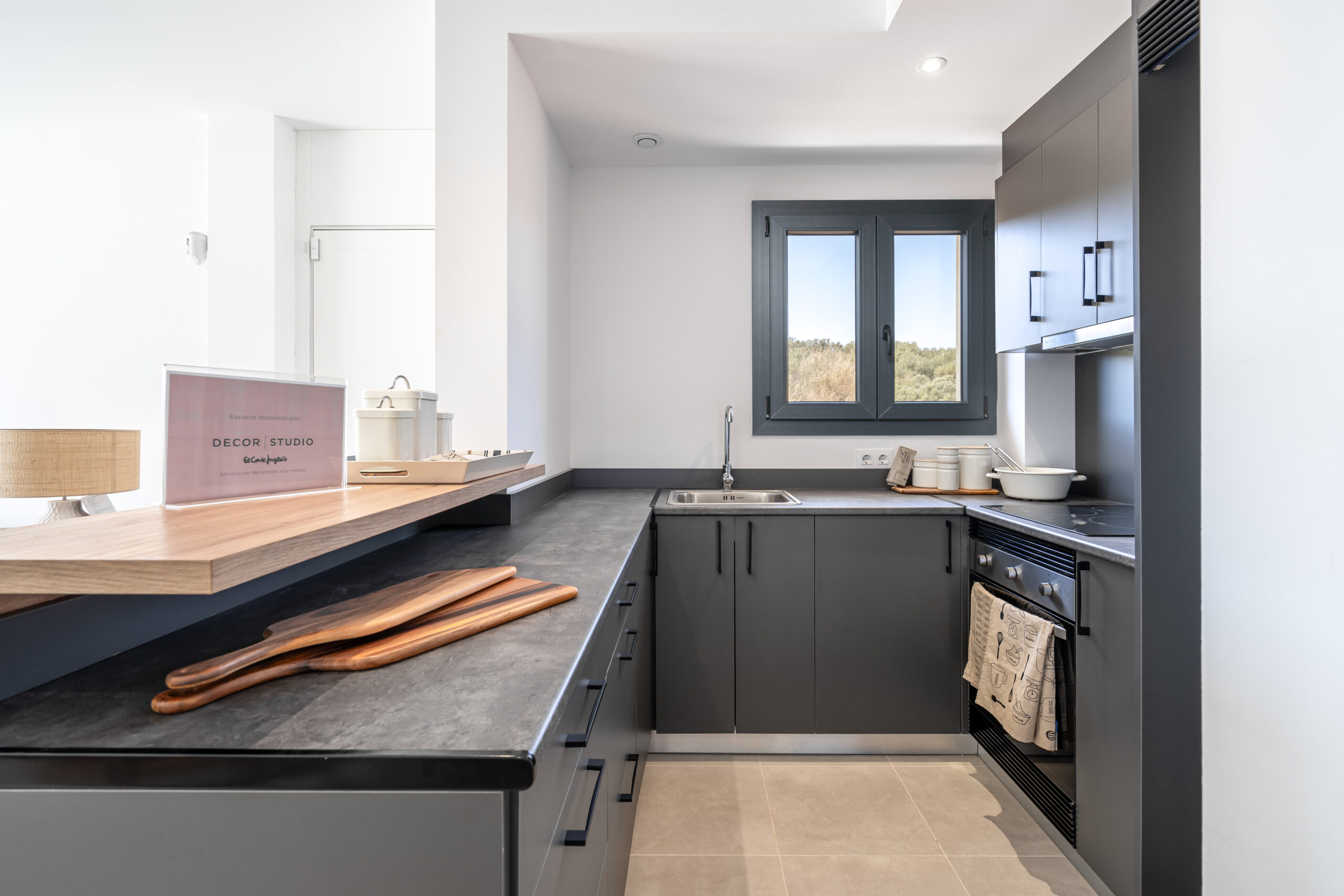 Moderne, kompakte Einbauküche in L-Form mit dunkelgrauen Fronten, Holzdetails und Blick aus dem Fenster auf die mediterrane Landschaft.