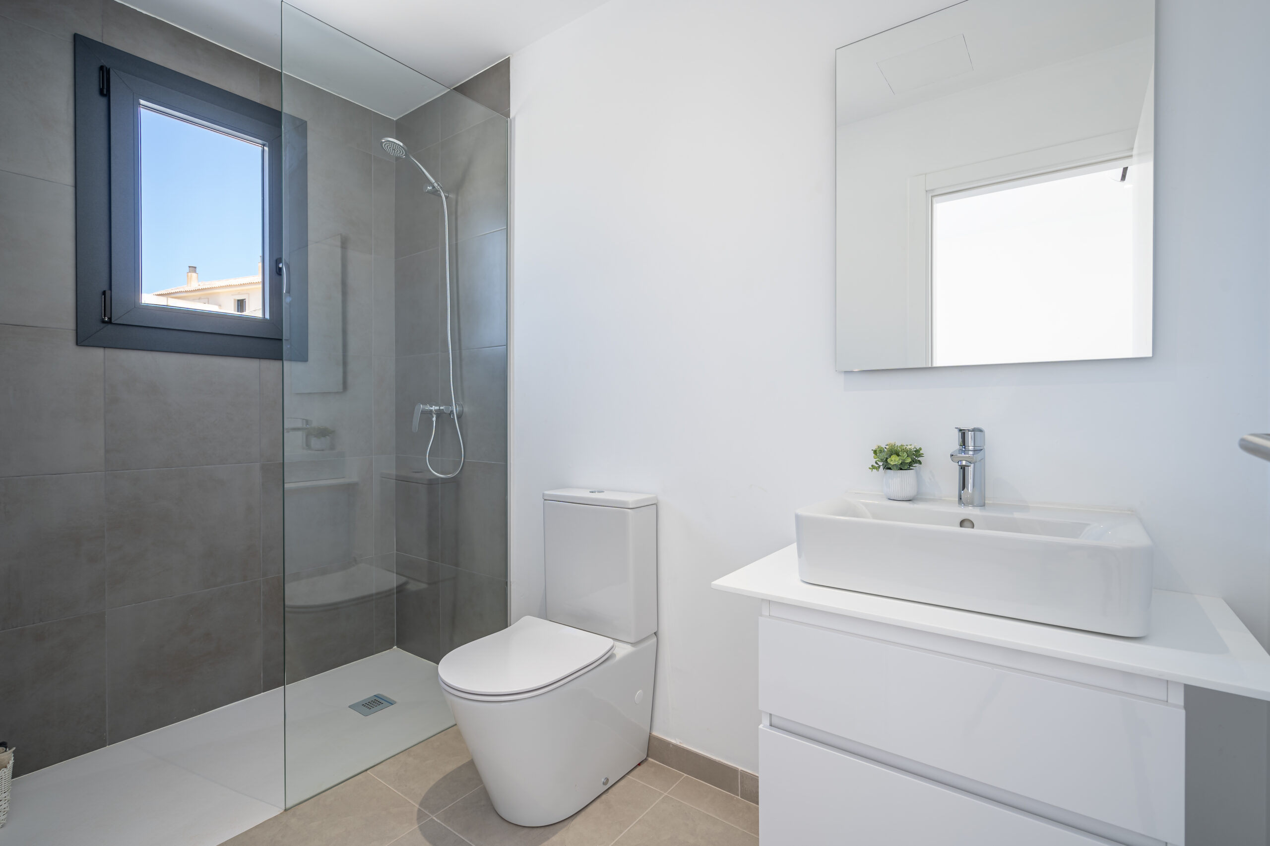 Modernes Badezimmer mit begehbarer Dusche, grauen Wandfliesen, weißem Waschtisch mit Spiegel und Fenster, das Tageslicht hereinlässt.